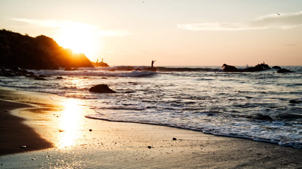 Fisherman stand on the rocks to cast their line as the setting sunlight streams across the shore and begins to set behind the cliff of La Punta.