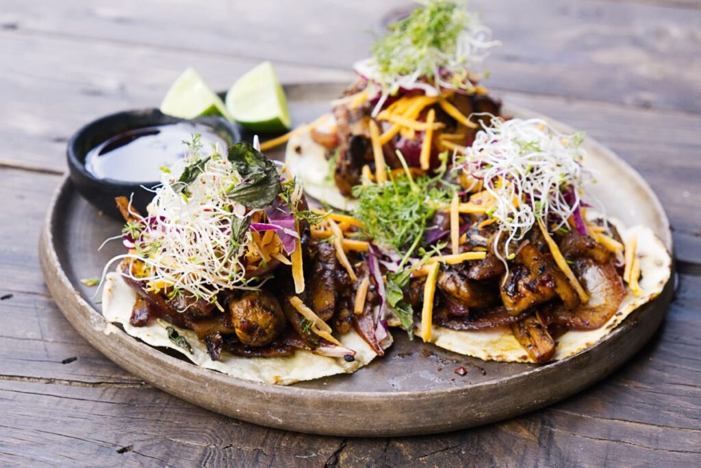 At Casa Armadillo Restaurant, Oaxaca are three mushroom tacos topped with veggies and fresh sprouts. They are presented on a round clay plate with a small bowl of salsa and two lime wedges.