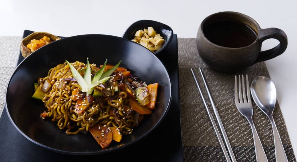 Dururu, a Korean restaurant in Oaxaca, serves their dishes on black plates with two small samples of kimchi. In the bowl is a ramen dish with beef and vegetables. On the side is a cup of tea in a clay mug and silver chopsticks, spoon, and fork.