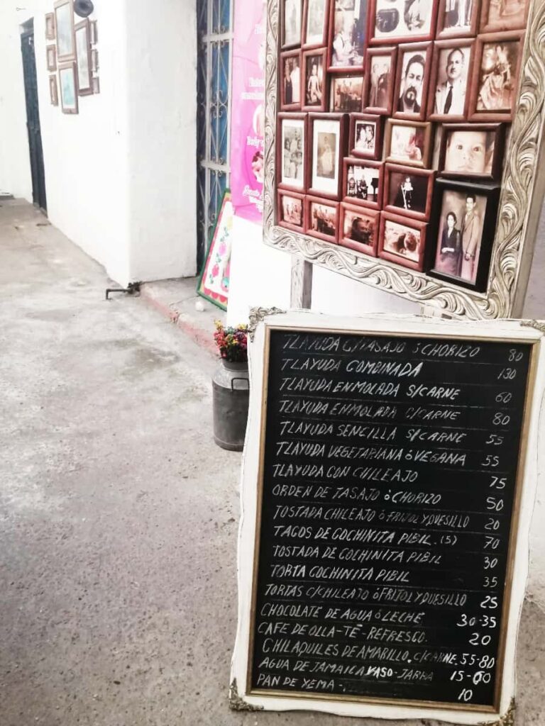At one of the best restaurants in Oaxaca for tlayudas, a handwritten chalkboard menu sits on the floor in front of a collage of family photos.