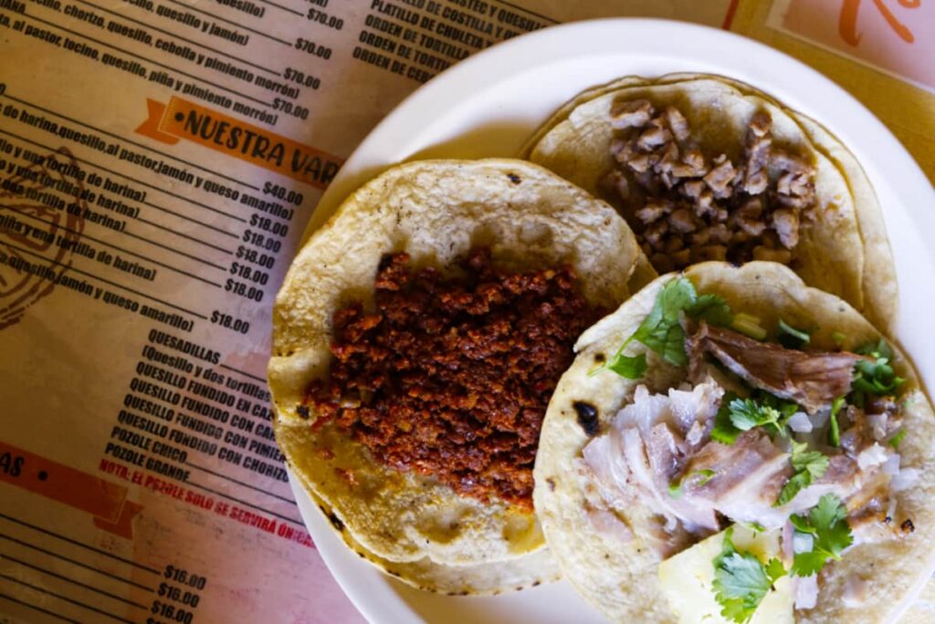 From above, a plate of three different tacos in Oaxaca - pastor, chorizo, and bistec sits on top of the menu at Taquería Tacomer.