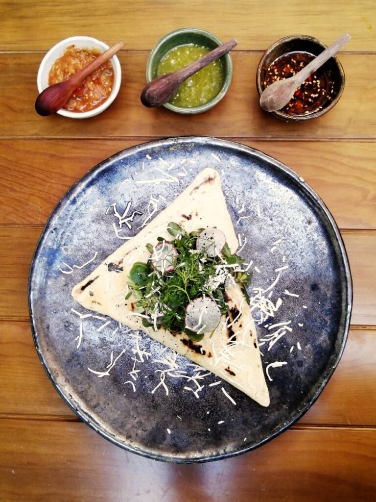 At Tizne, one of the best restaurants in Oaxaca, a view from above, a large triangular corn dough is plated topped with greens and served with a trio of salsas.