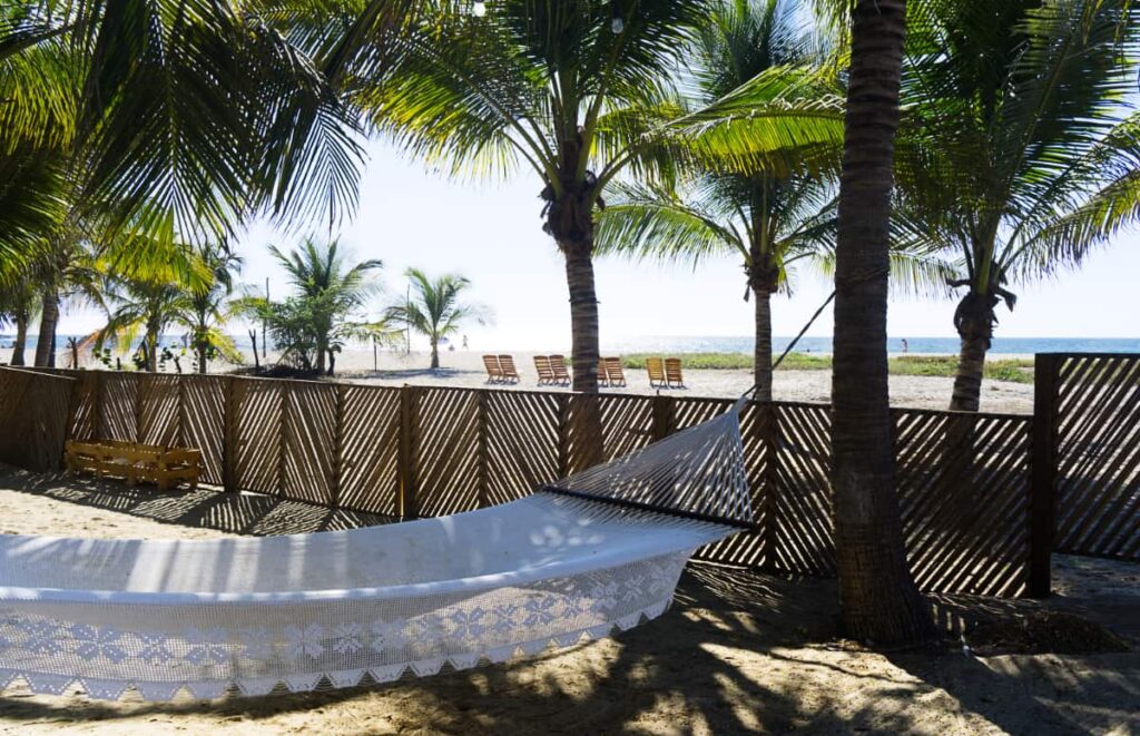 A white hammock with embroidered trim hangs under the palm trees. In the background is the ocean at La Punta with wooden beach chairs