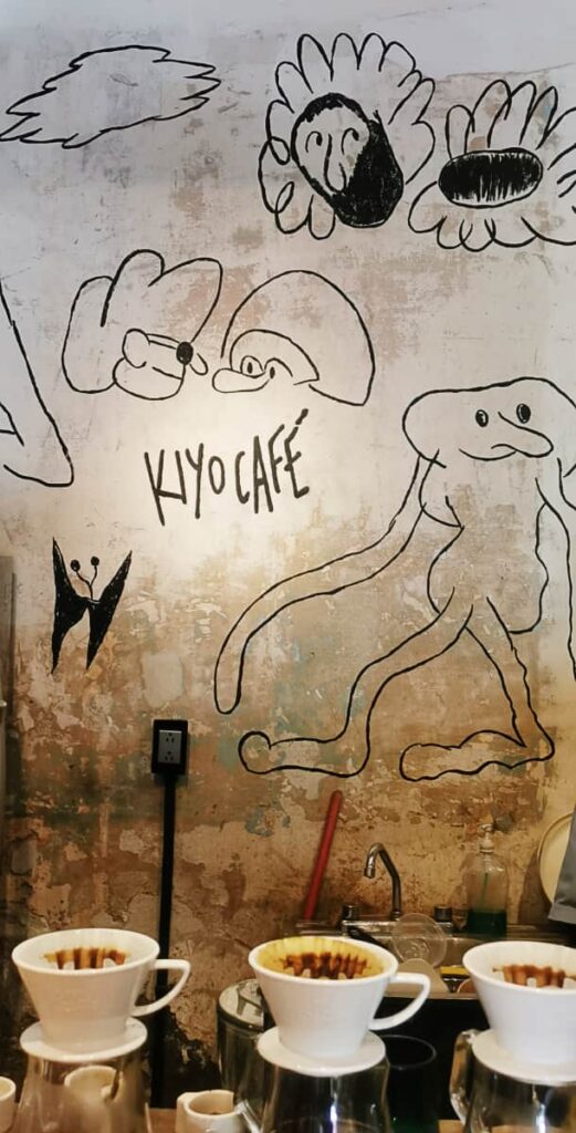 A black and white cartoon like drawing is painted on the back wall of Kiyo Cafe in Oaxaca. On the counter in front are several drip coffee filters on top of jars.