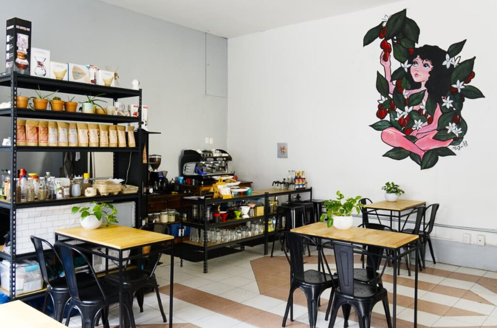 At Cafe SL28, a mural depicts a young girl sitting with her legs crossed amongst a plant as she picks a bright red fruit. Inside the cafe are tables and chairs as well as a shelf full of coffee products. Behind is the main bar with espresso machine and glasses.