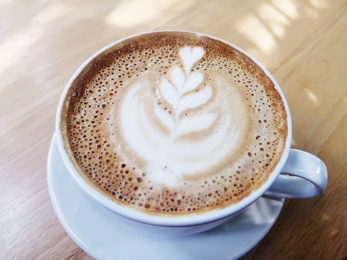 A cappuccino in a white mug sits on a wooden table. The coffee drink has lots of bubbles and a heart decoration drawn in the foam.