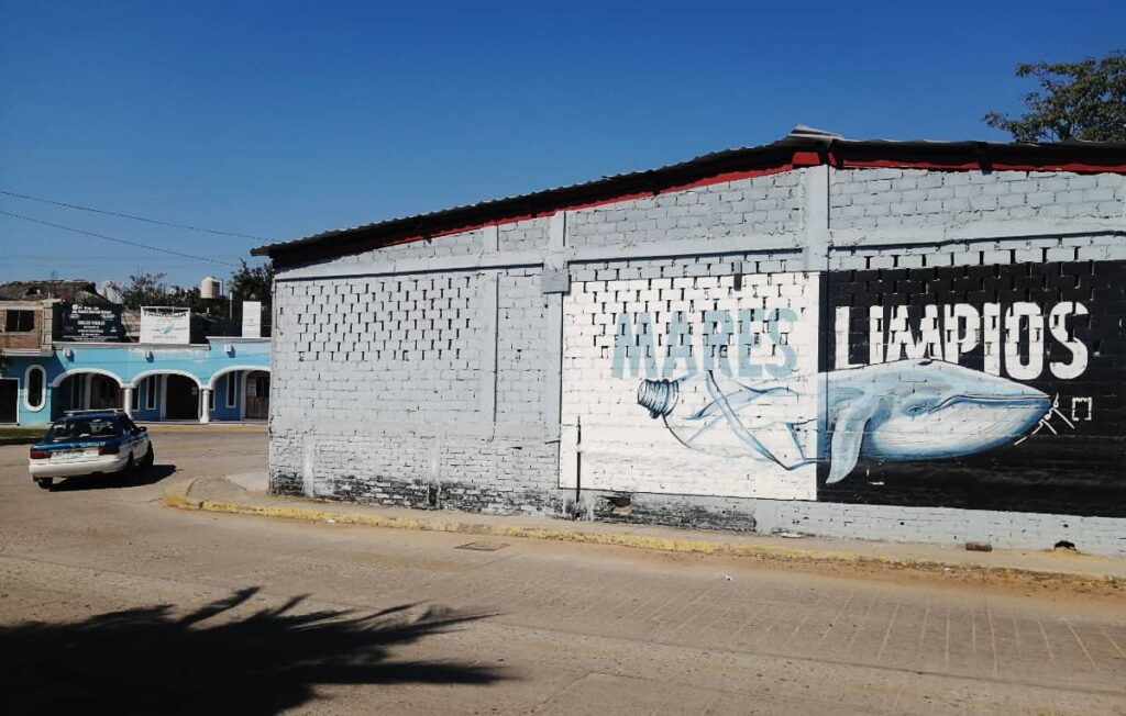 A street view of a Puerto Escondido neighborhood show a building with street art. It depicts a half whale, half plastic bottle with the words Mares Limpios which means Clean Oceans.
