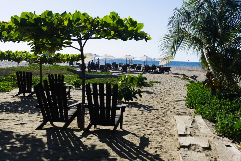 In La Punta, wooden Adirondack chairs sit in the sand facing the ocean under the shade of small green trees.