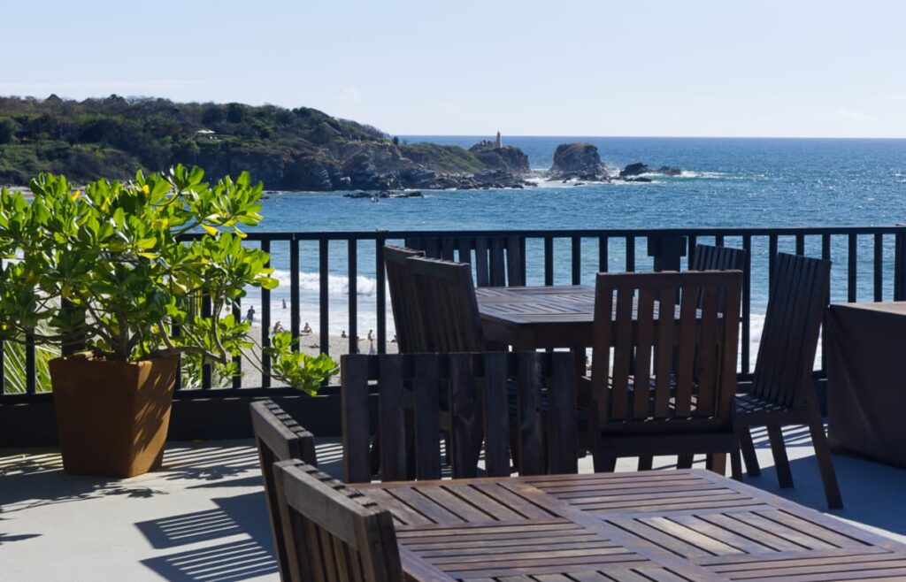 At Punta Zicatela Hotel, one of the best places to stay in Puerto Escondido, the rocky point of La Punta can be seen from the rooftop. Wooden tables and chairs sit in the sun with the ocean in the background.