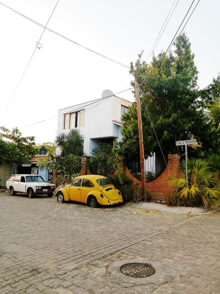 In the Puerto Escondido neighborhood of Rinconada, a yellow VW bug and an older white car are parked on the street. Behind them is a white house.