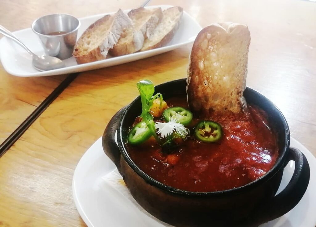 A bowl of red sauce with eggs and topped with sliced jalepeno and edible flowers. A piece of bread still out of the clay bowl. In the background are several more slices of bread on a plate with a side of salsa.