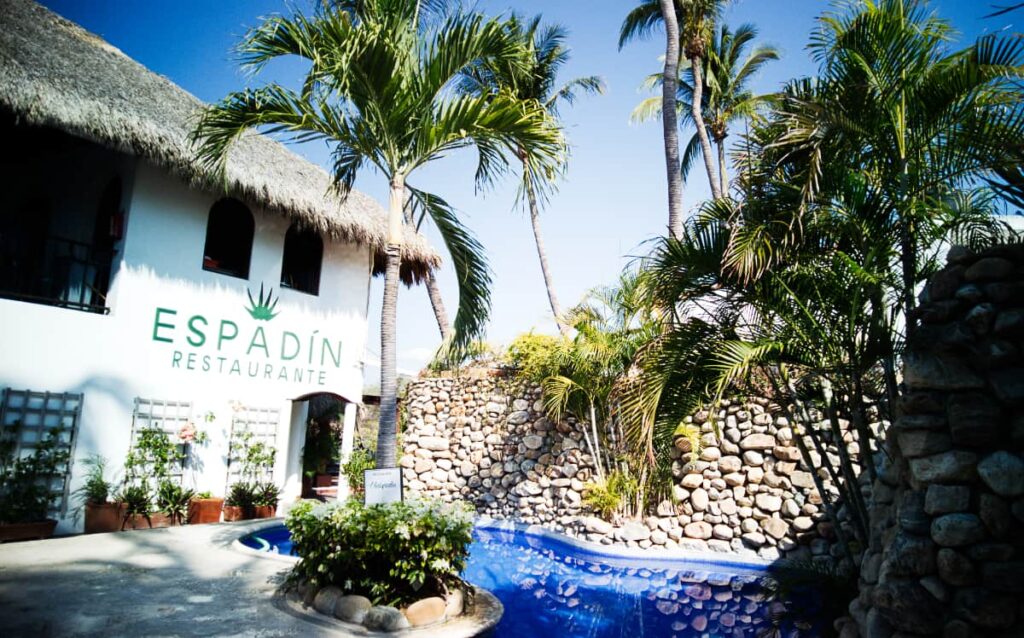 A small pool with curved edges is surrounded by palm trees and a white building with the words Espadin Restaurante at Villas Carrizalillo.