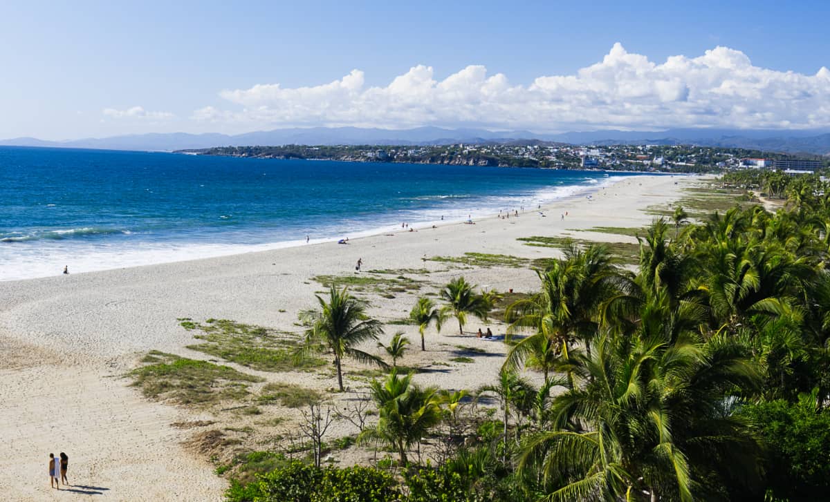 If you're wondering where to stay in Puerto Escondido, this overall, wide shot of people walking on the beach, palm trees, and the city in the background should give you a good idea.