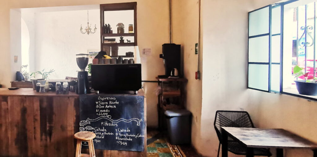 On top of the coffee bar at Filemon Cafe, Oaxaca is an espresso machine, coffee grinder, and mugs. On the right is a table and chair sitting under and open window with a plant.