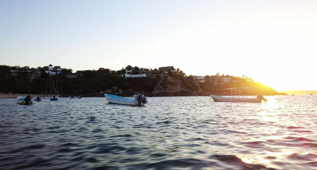 Boats are anchored in the bay near Puerto Escondido during whale season. The sun is starting to rise above the cliffside.