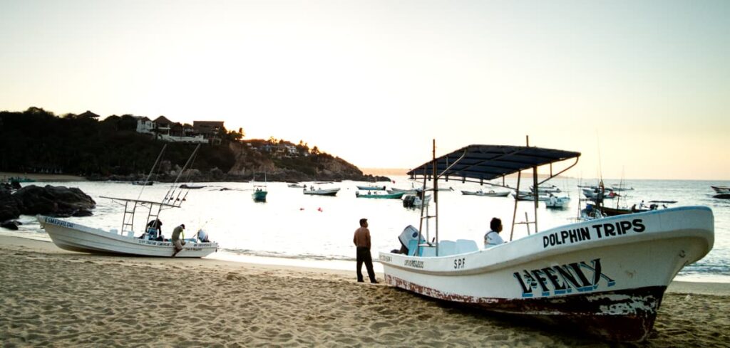 Two boats sit in the sand at this bay in Puerto Escondido during whale season. One boat that says dolphin trips on the side is preparing for a dolphin tour. In the background are other boats anchored in the bay.