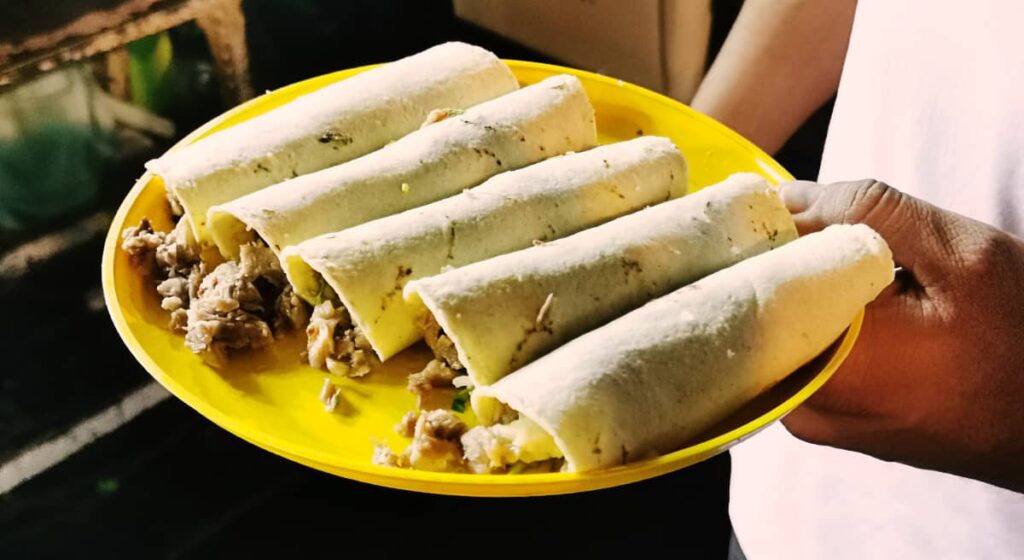 A man holds a plastic yellow plate of five Oaxaca tacos that are filled with meat and rolled.