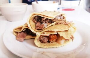 Three birria tacos are stacked on a white plate with a bowl of consume in the background.