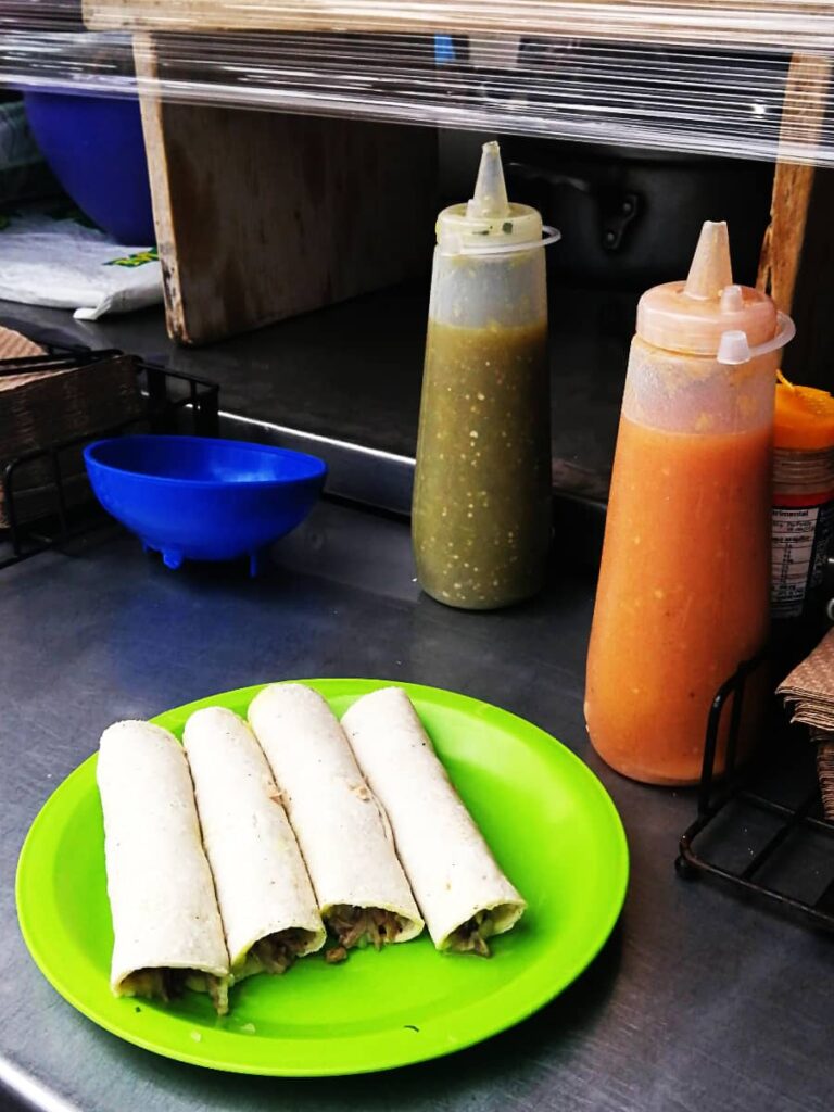 A Chefinita, a plate of 4 tacos are rolled up on a green plastic plate. Behind are two salsas - green and a very spicy red salsa for which these Oaxaca tacos are famous.