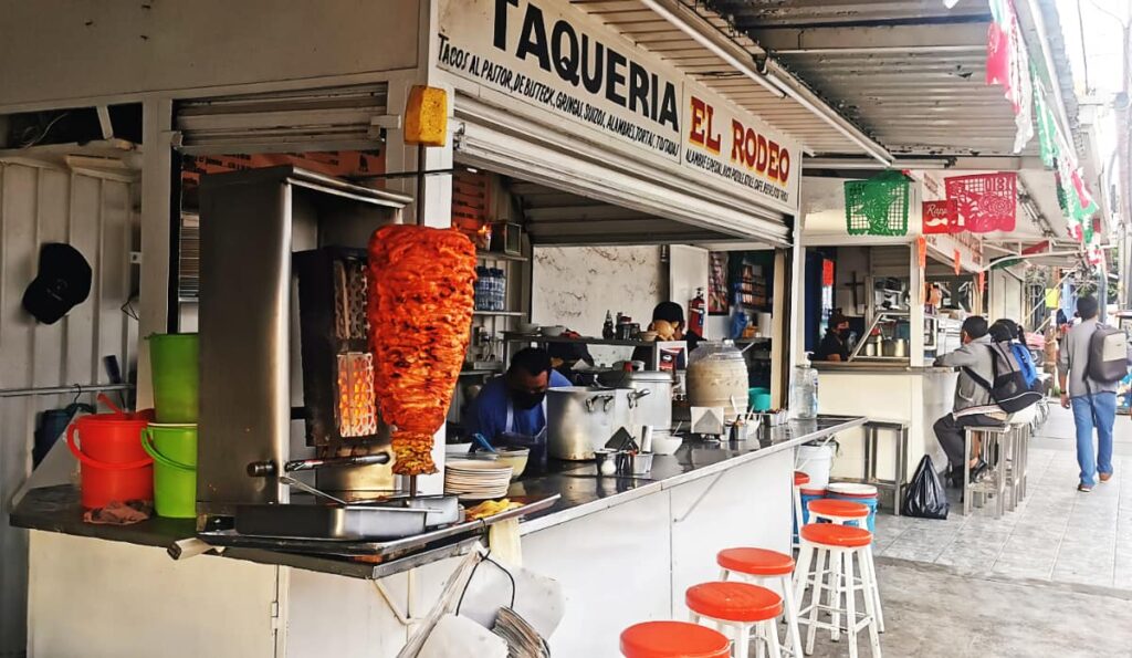 A rotating spit of tacos al pastor sits on the corner of this Oaxaca taco stand. Painted across the top is "Taqueria El Rodeo" in blue and red while a line of stools are in front. In the background are other taco stands.
