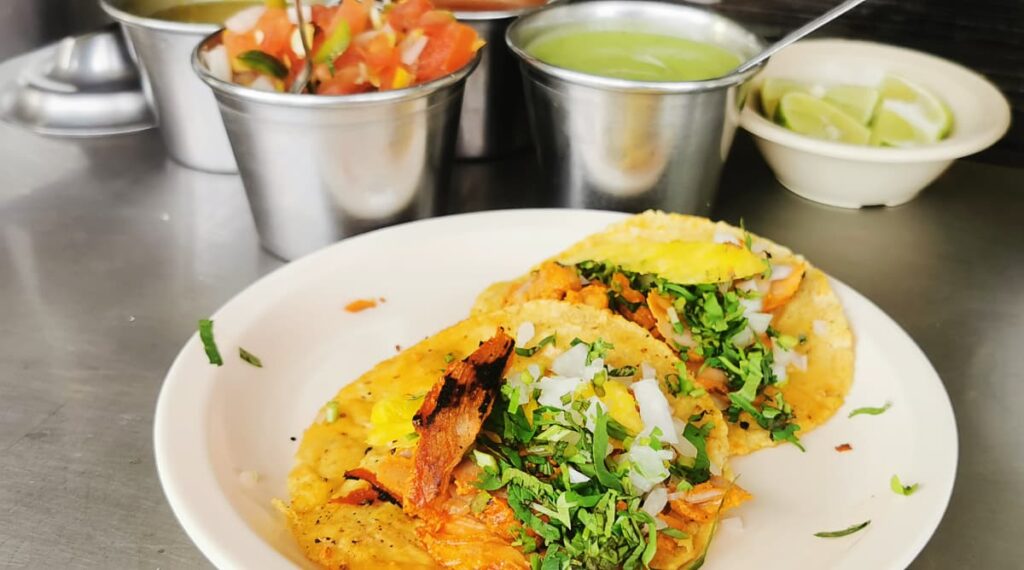 A plate of some of the best tacos al pastor in Oaxaca. The tacos are filled with pastor meat that is slightly burnt from the flame, pineapple, onions, and cilantro. In the background are several salsas and a bowl of limes wedges.