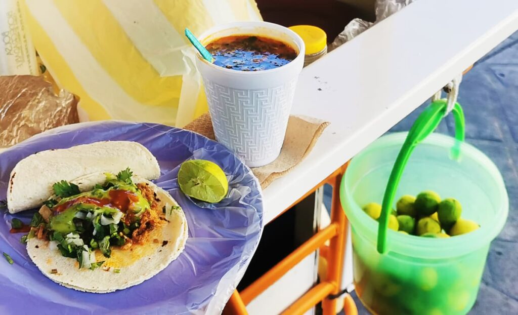 Two tacos de res and a cup of consume are balanced on the edge of a taco stand in Oaxaca. Below hangs a bucket of limes.