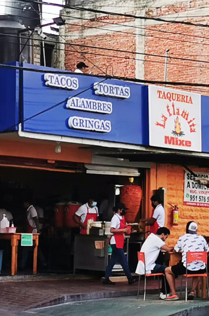 From the outside of Taqueria La Flamita Mixe, customers sit at an outdoor table while servers come from inside. A man stands near the front to tend to the tacos al pastor which are rotating on a spit in front of a flame. The sign above reads the name of the taqueria and the words tacos, tortas, alambres, gringas.
