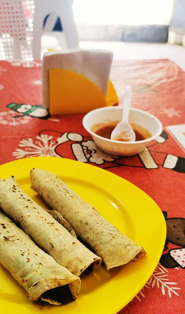 A yellow plate of three rolled tacos sits on a table with a red table cloth. In the background are napkins and a bowl of hot sauce.