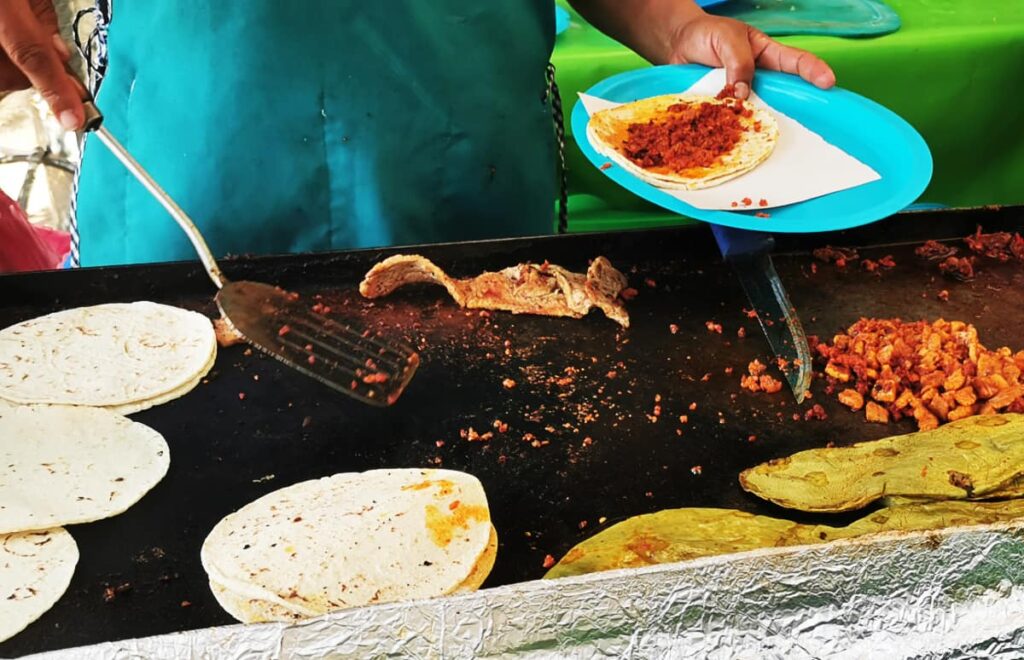 At a El Llano taco stand, a woman grills chorizo, nopales, and bistec while holding a blue plastic plate with a chorizo taco.