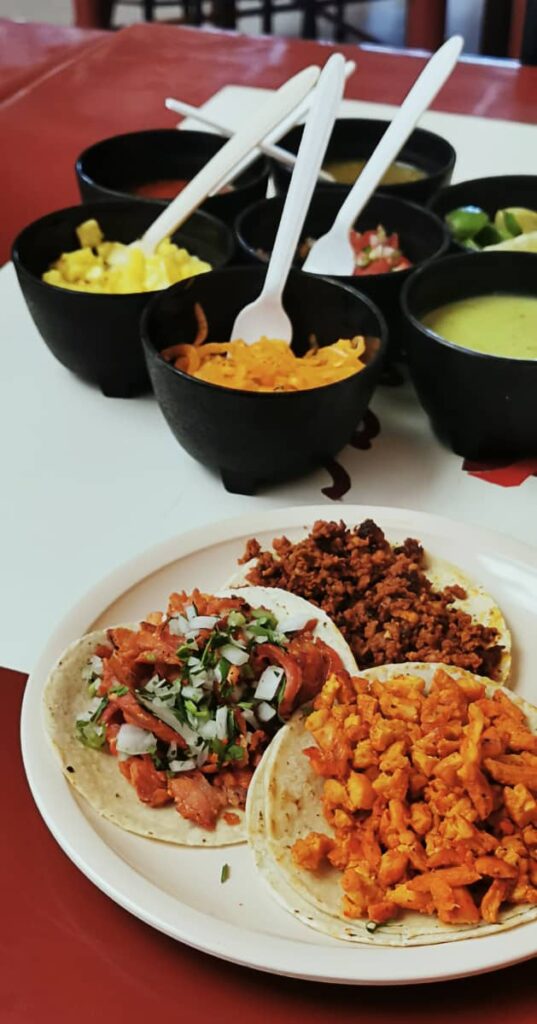 A plate of three tacos - al pastor, chorizo, and bistek sit on a table at Taqueria Los Combinados, the only place to eat Oaxaca tacos 24 hours per day. Behind are 7 bowls of salsas, hot peppers, and limes.