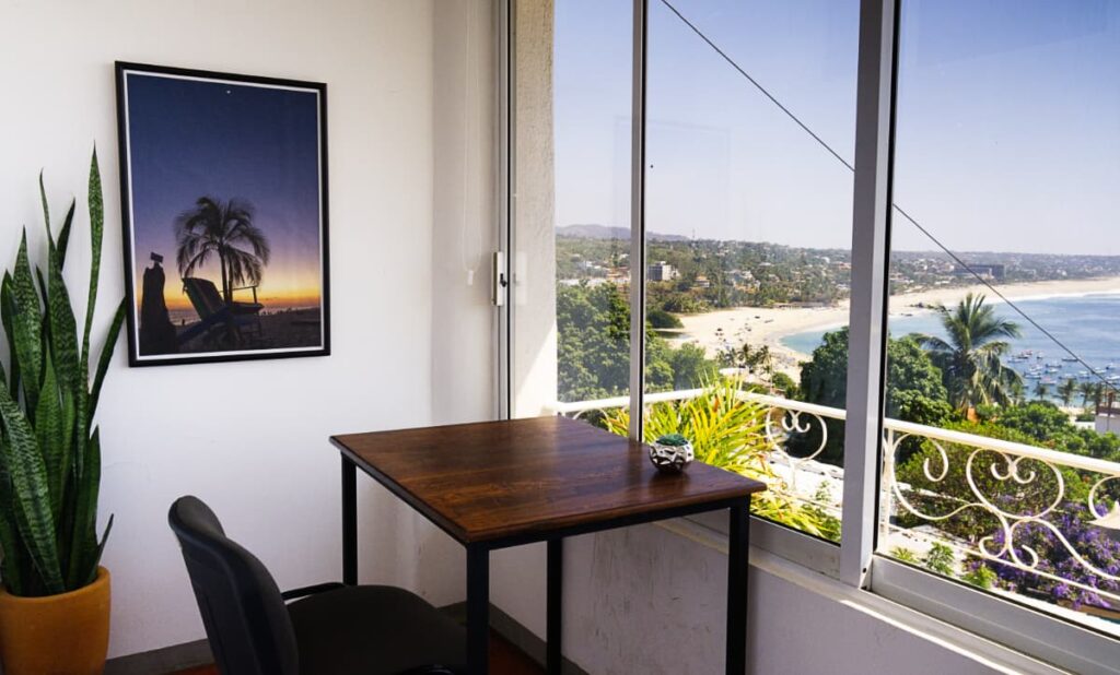 In the private office at 401 coworking space in Puerto Escondido, a wooden desk and black chair face a view of the ocean through the windows. On the wall is a sunset print of a palm tree and in the corner is a green plant.