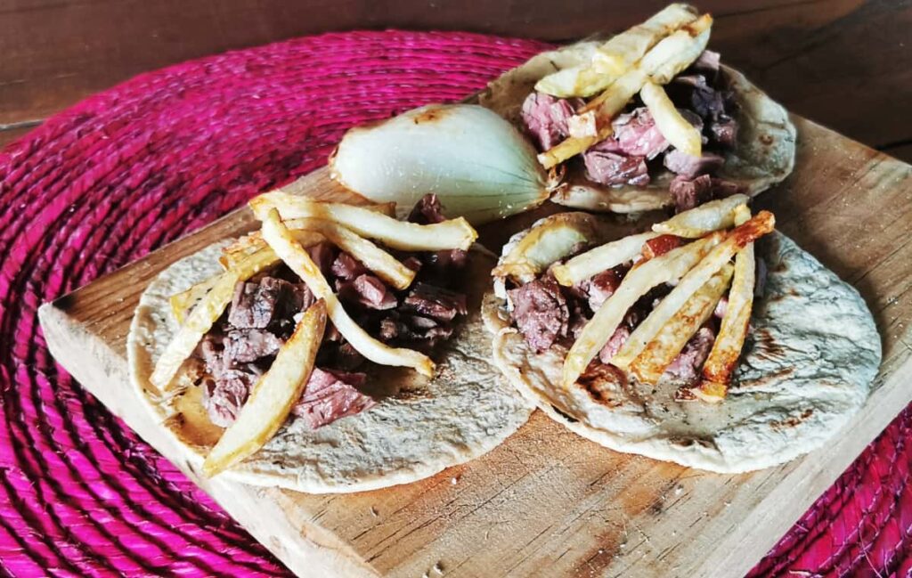 At one of the best restaurants in Puerto Escondido, three arrachera tacos topped with fries are served on a wooden plank that sits on a magenta woven place mat. On the side is a half grilled onion.