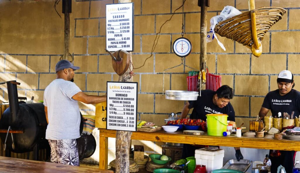 Two men work behind the counter at Lenos & Carbon, one of the best places for barbacoa in Puerto Escondido. Above is a scale to weigh the barbacoa meat and on the left is a grill to cook the costillas that are sold on Saturdays.