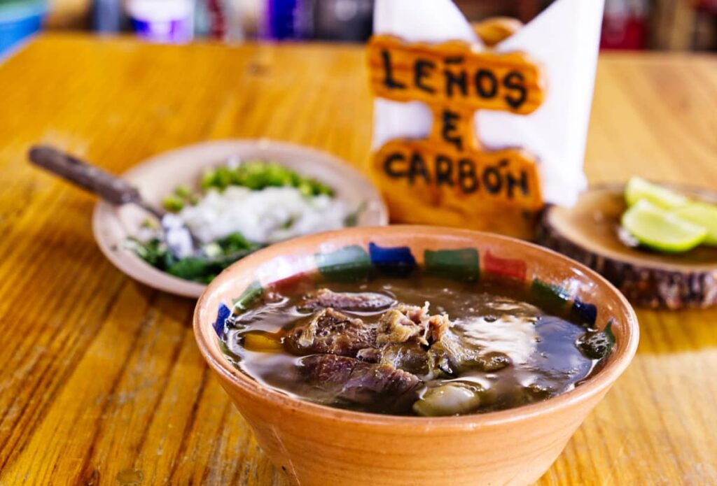 A bowl of broth and barbacoa de borrego, or lamb barbeque sits on the table at Lenos & Carbon. Behind is a plate of cilantro, onions, and jalapeno as well as a plate of limes.