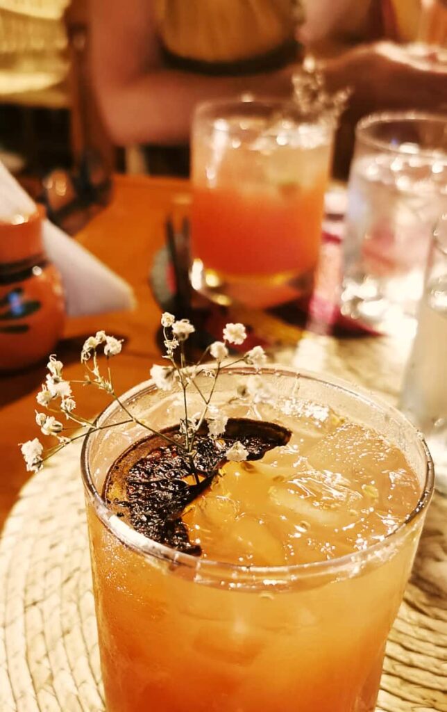 On the table at this restaurant is a mezcal craft cocktail served in a glass with a slice of burnt orange and small white flowers.
