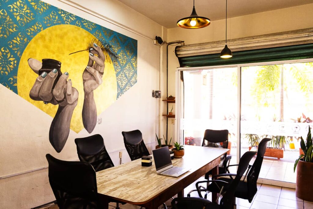 A long table surrounded by black office chairs sit in front of a large sliding door at Losodeli's formal coworking space in Puerto Escondido. On the wall is a large graphic art piece depicting two hands and forearms holding a feather quill and ink well on a golden and turquoise background.