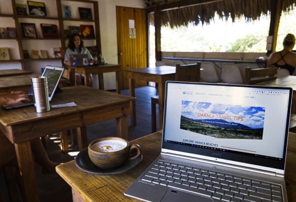 At Malagua coworking in Puerto Escondido, are several wooden tables, one with a cappuccino next to my computer showing the main page of Oaxaca Travel Tips. In the background several digital nomads are working from their computers at different wooden tables.