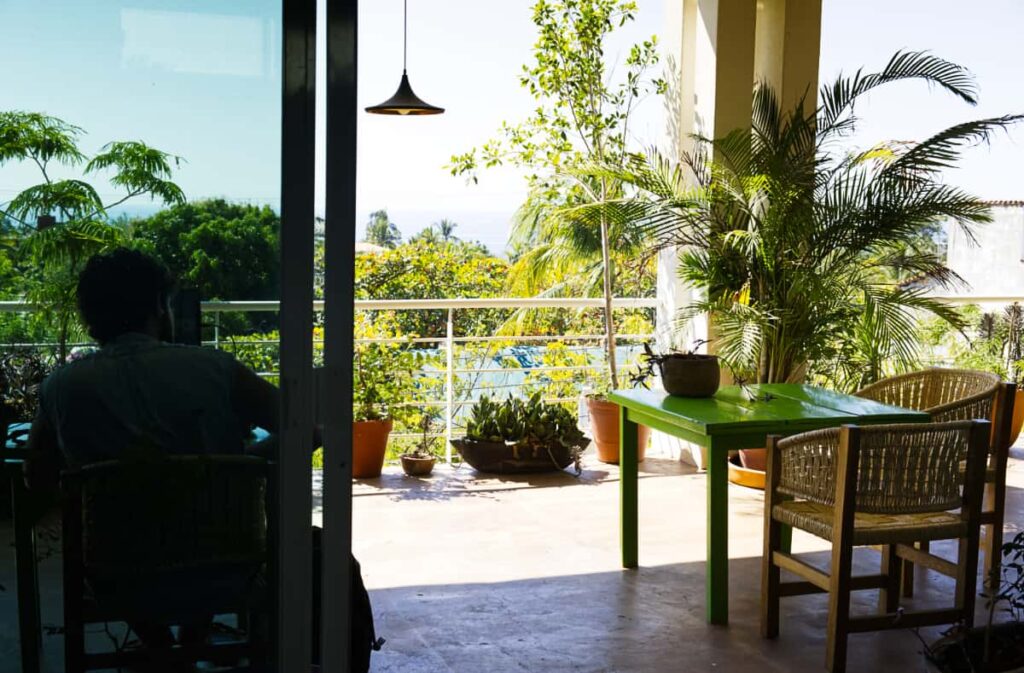 A man is silhouetted sitting in front of the sliding glass door on the balcony of nuu coworking in Rinconada, Puerto Escondido. Other wooden tables and plants are on the balcony which has a distant ocean view.
