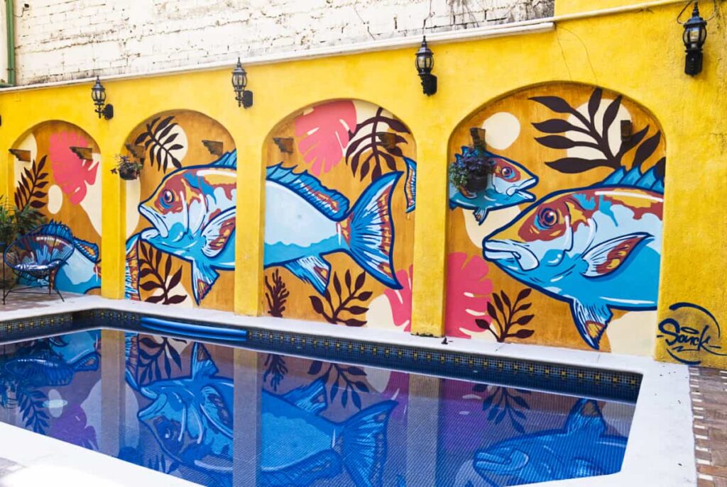 The blue pool at Pargos Hotel and coworking is decorated with a graphic fish art painted on the yellow wall behind the pool.