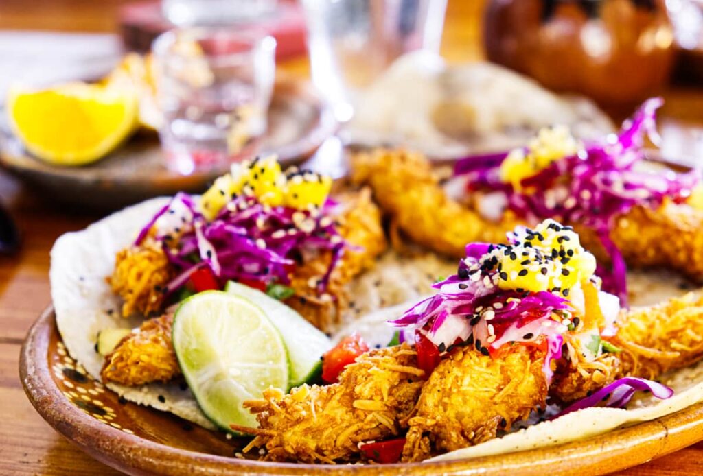 Restaurant Rey Shaman serves crispy coconut shrimp tacos topped with a red cabbage slaw and fresh fruit. In the background is a shot of mezcal with citrus slices.