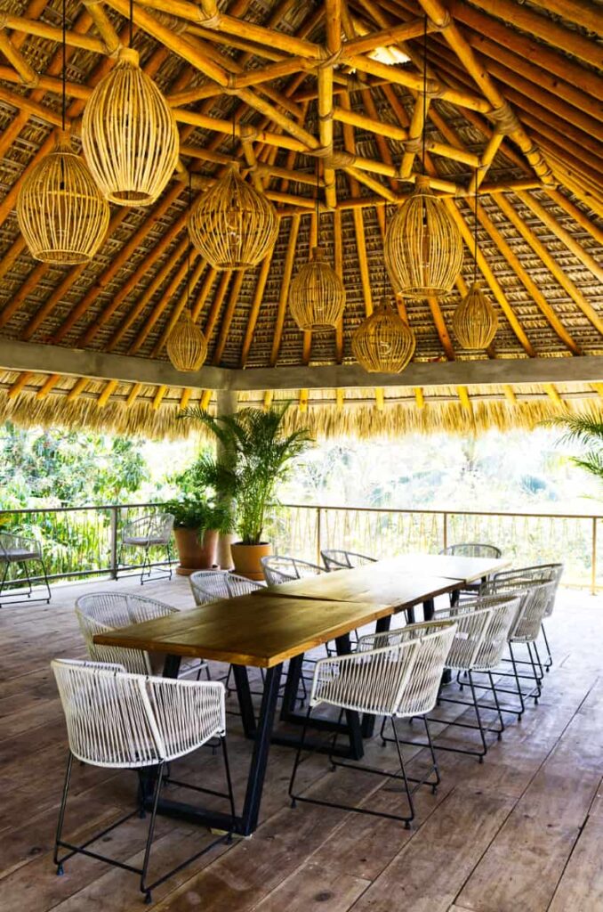 The coworking space at Raiz Boutique Hotel features a long wood table surrounded by white chairs. Several ratan light fixtures hang from the palapa roof above.