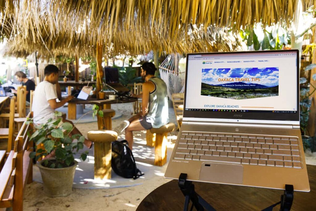 Digital nomads sit on stool under the palapa tables at Sabina coworking in La Punta, Puerto Escondido. In the foreground is my computer on a laptop stand, showing the main page of Oaxaca Travel Tips.