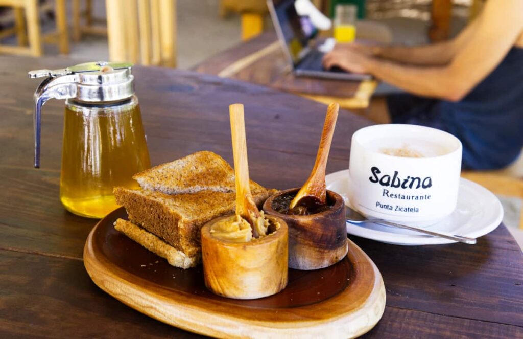 Next to a cappuccino sits stack of toast sits on a wooden plate with a wooden bowl of peanut butter, another of jelly, and a canister of honey. In the blurred background, a digital nomad works from his laptop while drinking an orange juice.