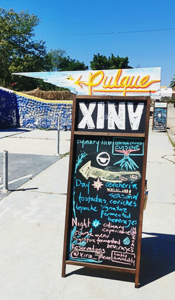The sign for Xina, a fine dining restaurant in Puerto Escondido tells passers about ceviche, tostadas, and signature fermented beverages. At night, their culinary experience is highlighted which includes a 5 course tasting menu with fermented beverages. At the top, a wooden sign is attached to advertise Pulque, a fermented beverage, in bright yellow letters.