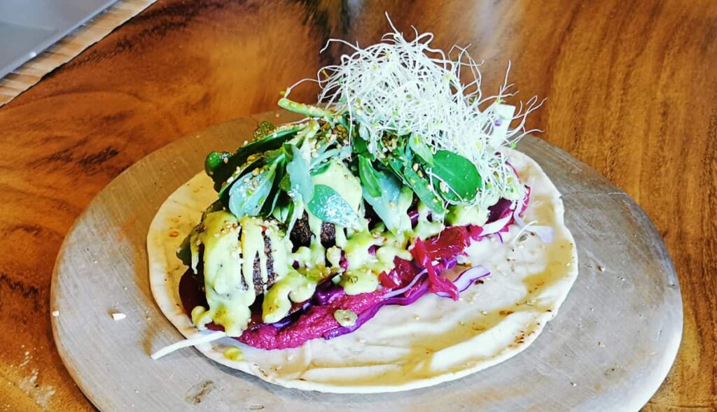 At Aguacate Veggie Bar, a vegetarian restaurant in Oaxaca City, an open faced falafel wrap sits on a flat plate on top of a wooden table. A salsa and fresh greens are loaded on top of the wrap.