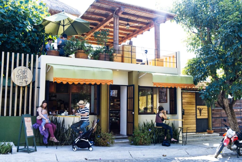 At Cafe Nopal, one of the best breakfast in Puerto Escondido, A couple with a baby sit at one small table outside of the entrance, while a single woman sits at another. On the upper level is the rooftop with tables and a roof for shade.