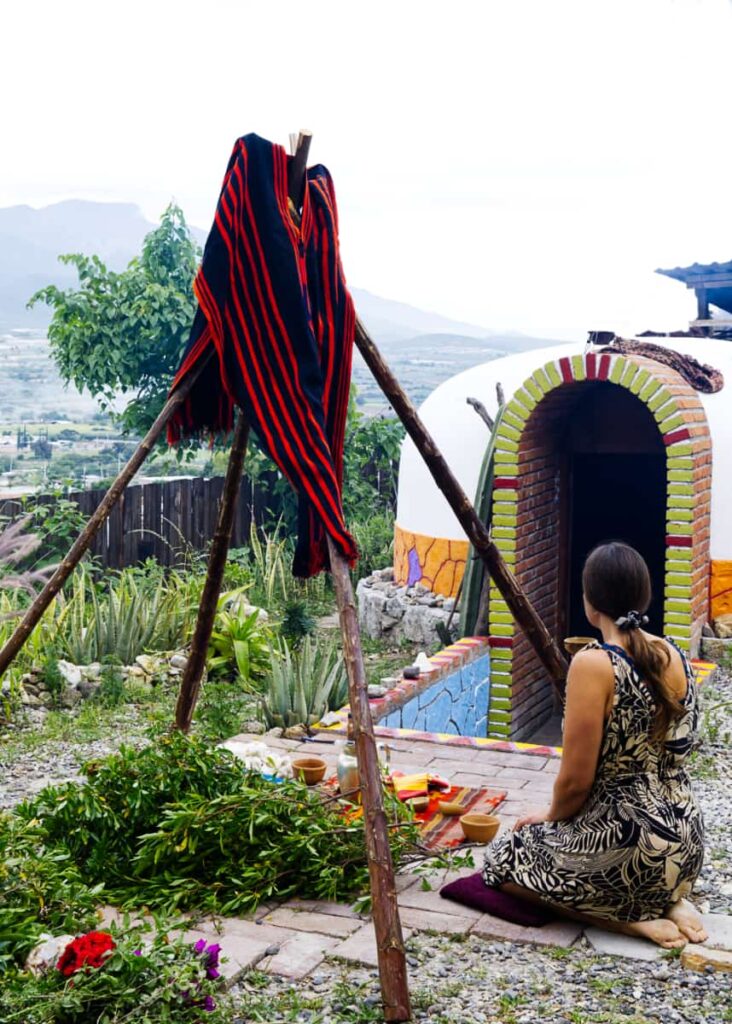 A woman sits in front of a temazcal in Oaxaca, overlooking the mountains. In front of her is a large piles of herbs and some small dishes as well as four sticks tied like a teepee with a red and black cloth.