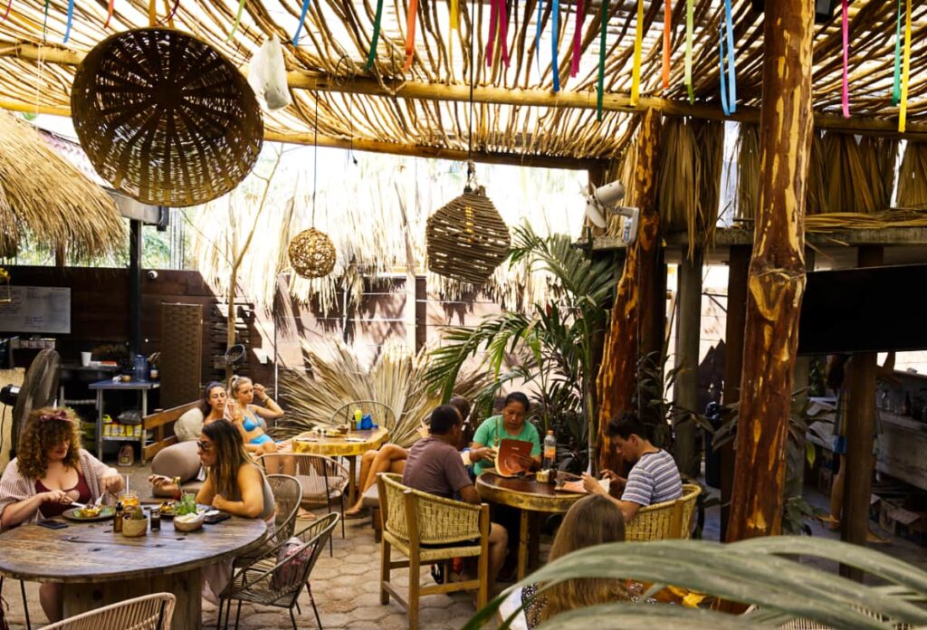 Several round tables are filled with groups of people eating brunch at Malagua Cafe in Puerto Escondido. Above are several woven light fixtures and a colorful strings hanging from the natural roof for decoration.