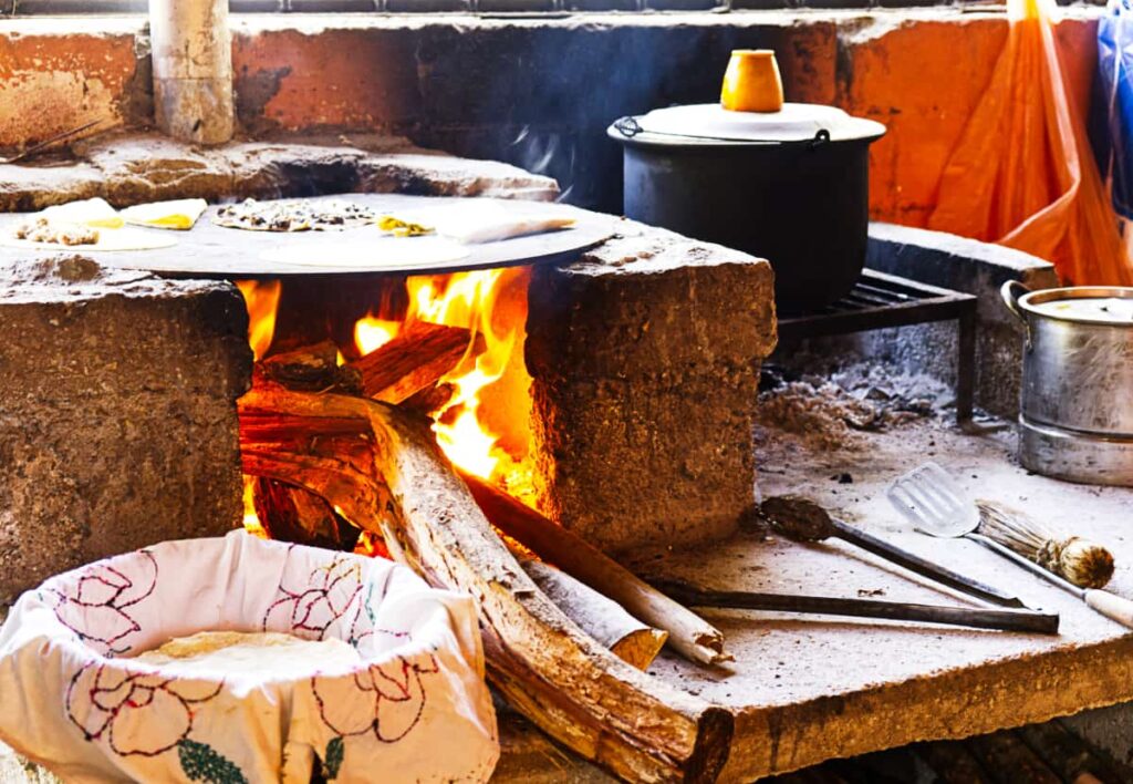 A wooden fire heats the comal with several breakfast foods cooking on top of the round metal surface. In front of the fire is a basket with tortillas inside.
