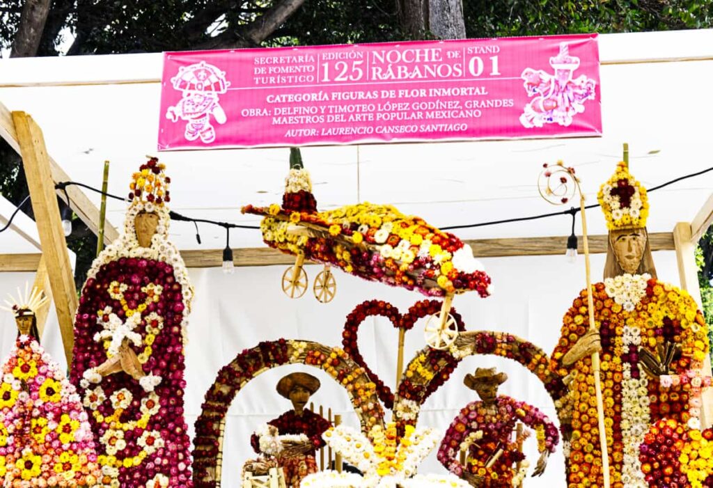 An entry in the immortal flower category of La Noche de los Rabanos competition in Oaxaca City depicts several biblical figures, an airplane, and a small Oaxacan scene. Small flowers are used to create the figurines which range in color from yellow to magenta with a splash of white.